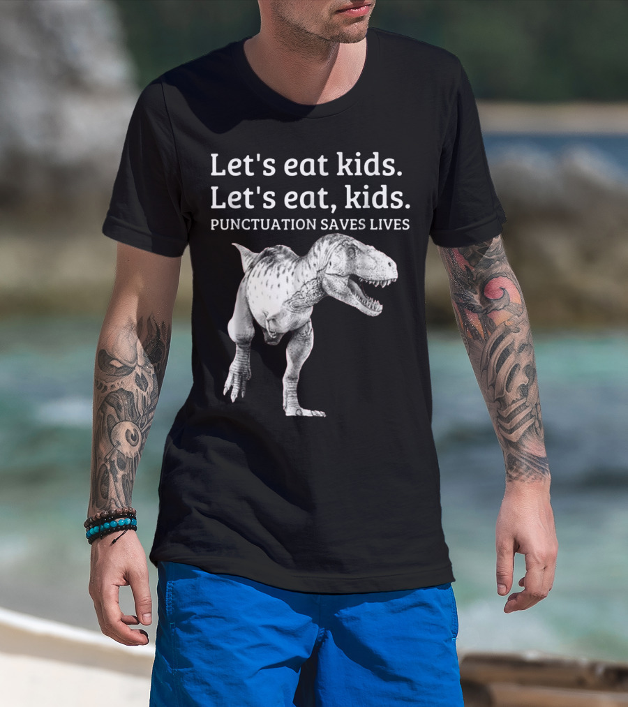 Man with tattoos models Let's Eat Kids Let's Eat Kids Punctuation Saves Lives Dinosaur T-Shirt at beach.